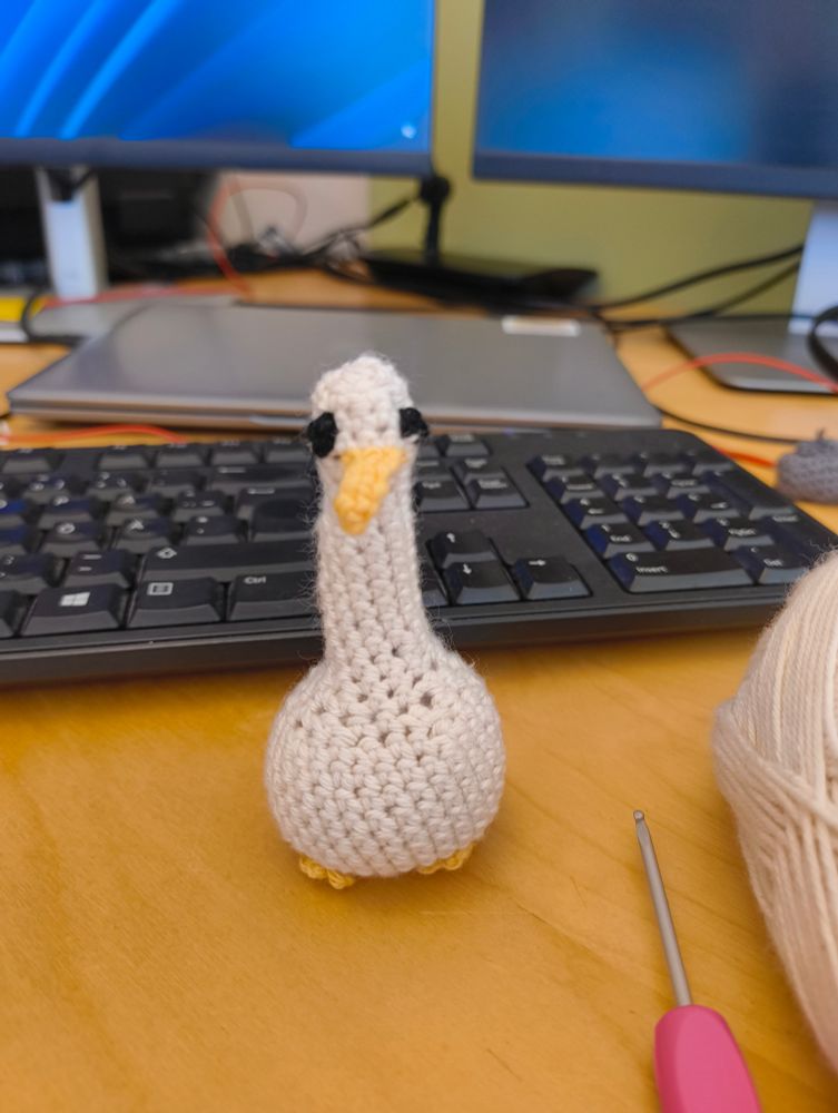 A white crocheted bird with a round body and long neck. The beak is yellow and the eyes are black. It's supposed to be a seagull but you may interpret it in any way you want to.