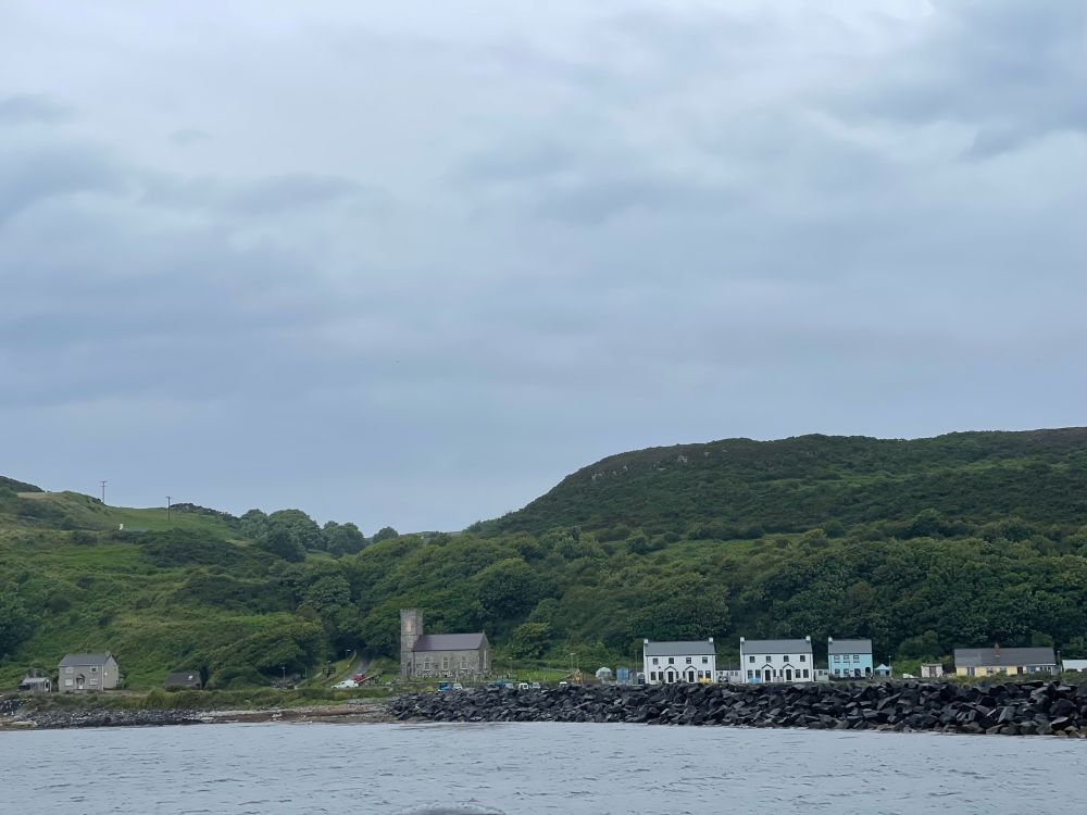 Looking back at the harbour on Rathlin Island from the ferry back to Ballycsstle. A stone church and some houses are on the seafront, nestled beneath some wooded hillside. 