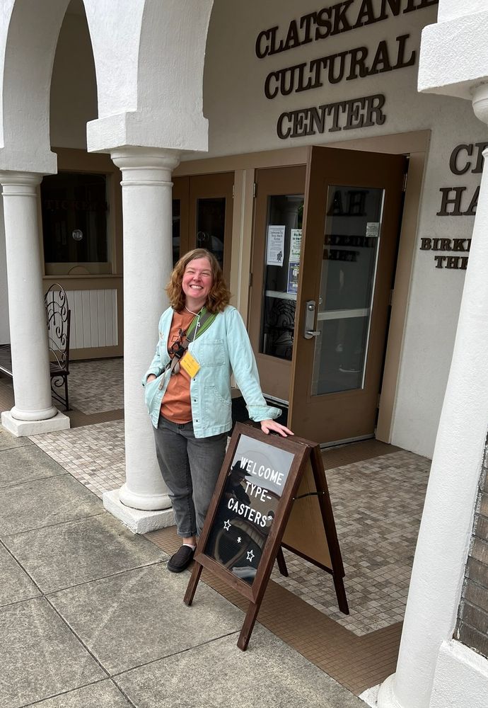 A smiling woman stands in front of the Clatskanie Cultural Center in Oregon, leaning on a sandwich board that says "Welcome Typecasters"
