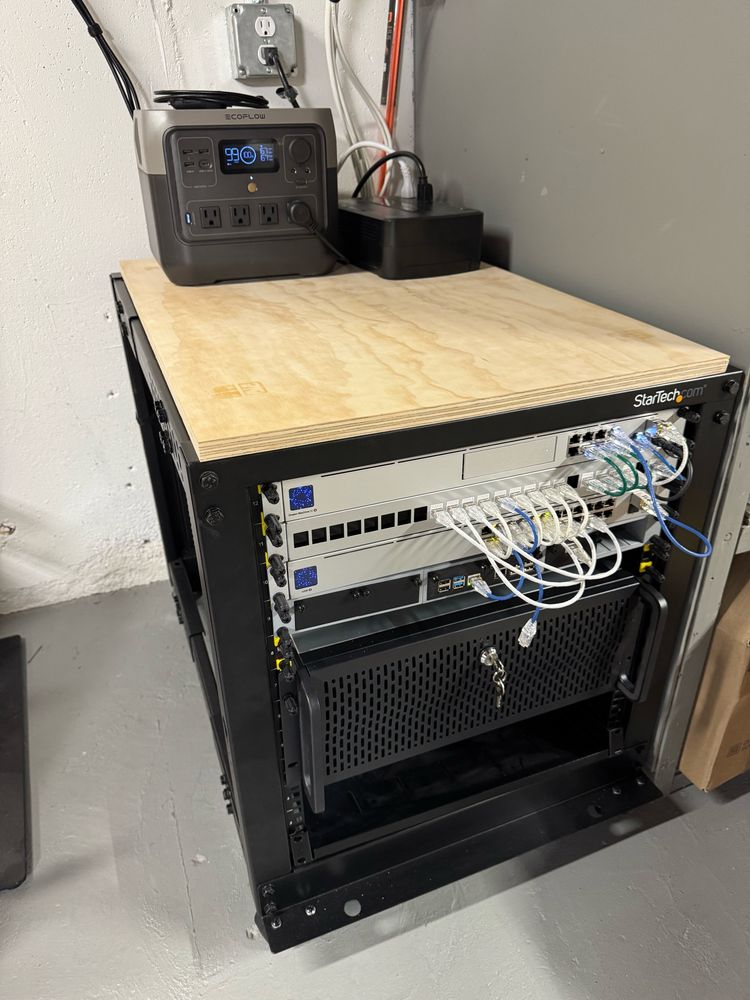 Small server rack with a plywood shelf on top. The shelf has an ecoflow battery and UPS connected together to power the equipment within the rack. 