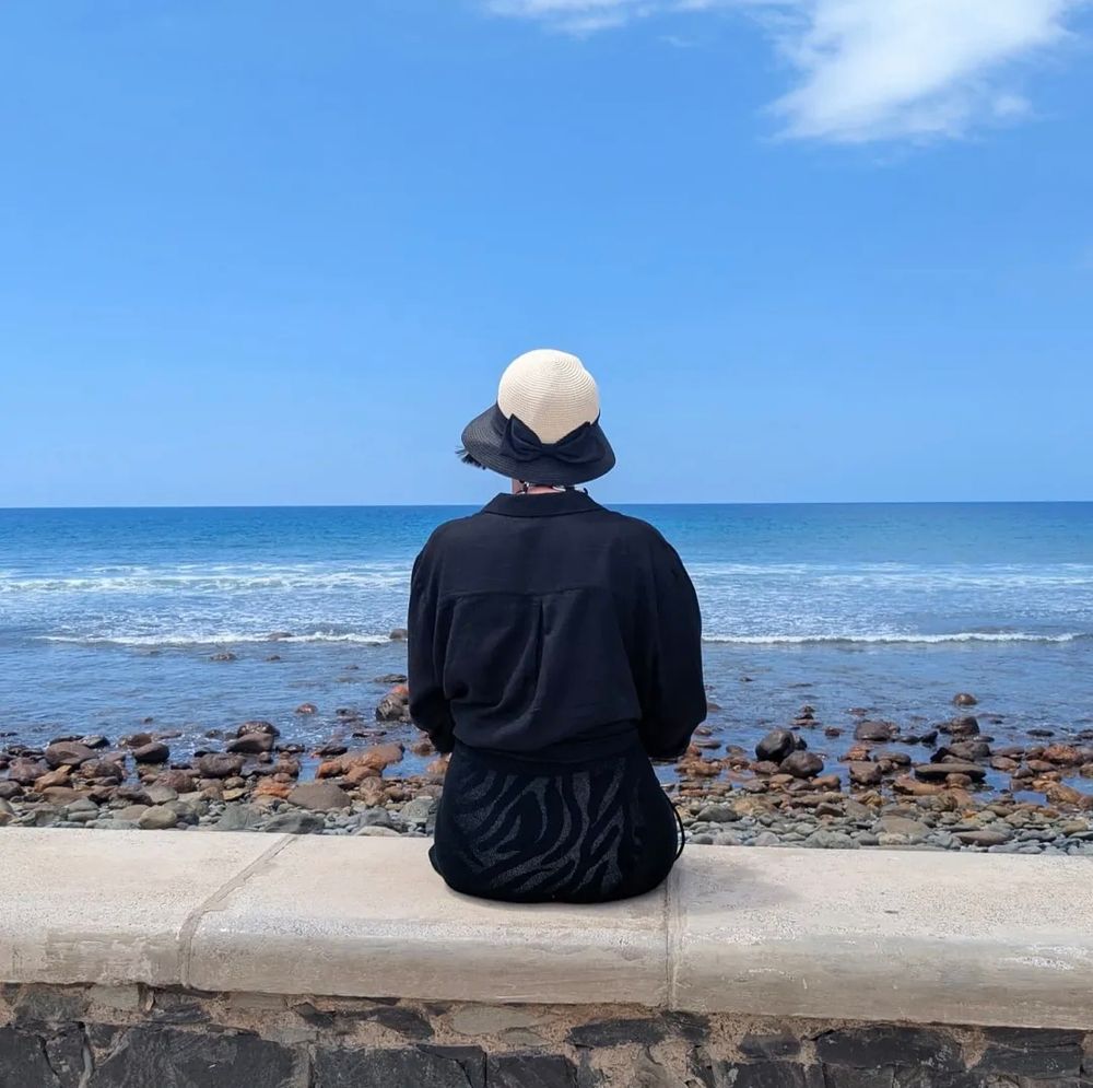 Woman with her back to the camera wearing a hat and looking out on a blue sea under a blue sky. 