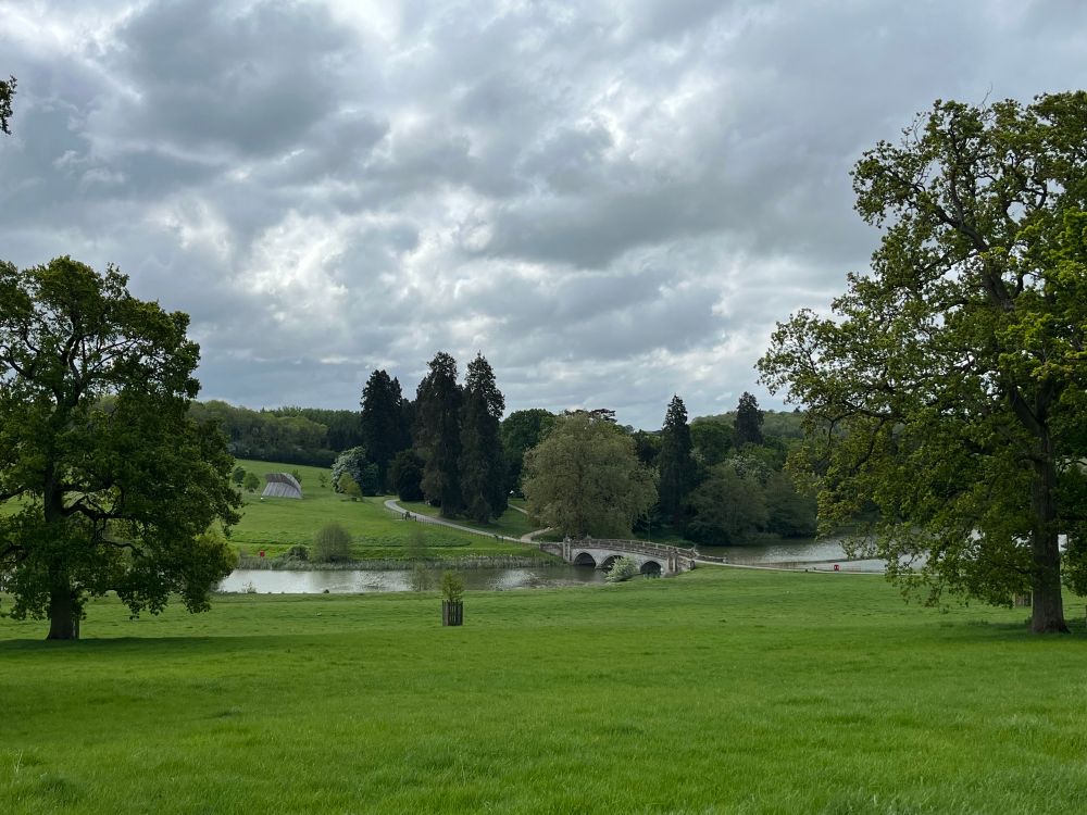 Grey clouds over a green landscape. Tall trees in the foreground. A stone bridge over a lake in the distance 