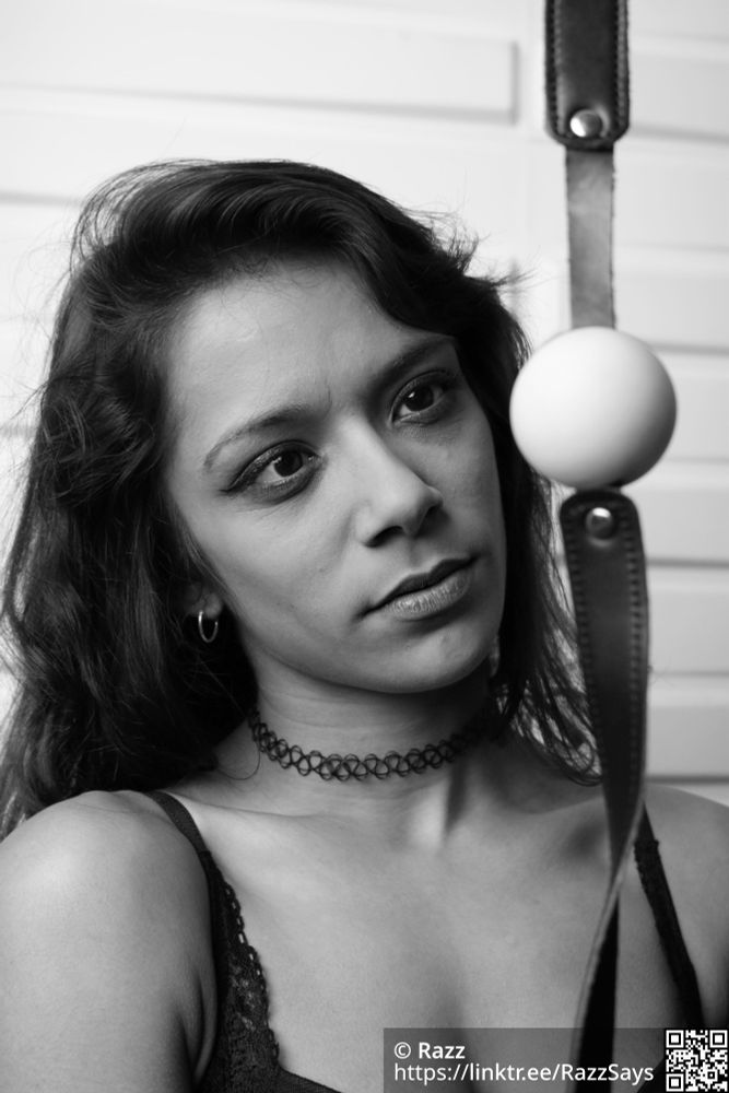 A mixed race woman looks at a ball gag with a "what?" expression on her chops.