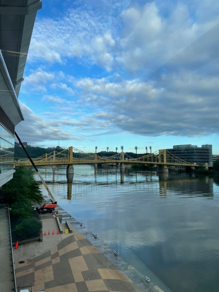 A view of the river from the Pittsburgh Convention Center, with a view of blue skies and white clouds reflected in the water, crossed by yellow bridges.