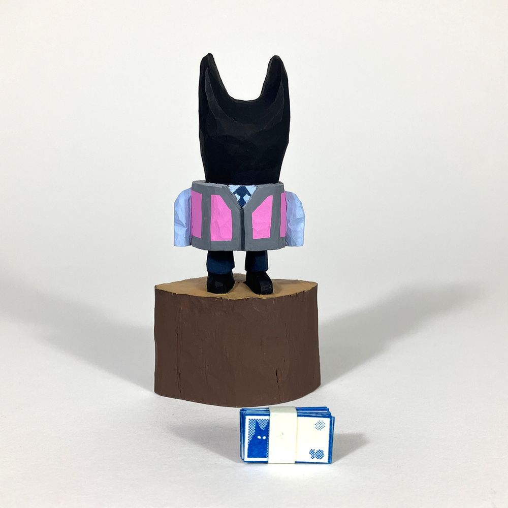 Cherry wood carved figure of a cat like humanoid form in a shirt and tie and a hi viz waistcoat. It’s stood on an uncarved section of the wood. A band of tiny paper printed banknotes is laying near the base of the figure