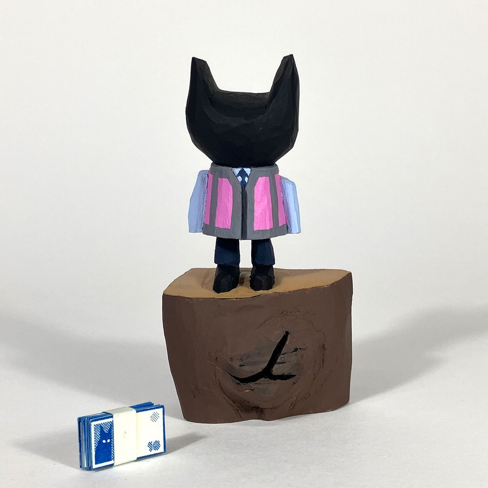 Cherry wood carved figure of a cat like humanoid form in a shirt and tie and a hi viz waistcoat. It’s stood on an uncarved section of the wood. A band of tiny paper printed banknotes is laying near the base of the figure