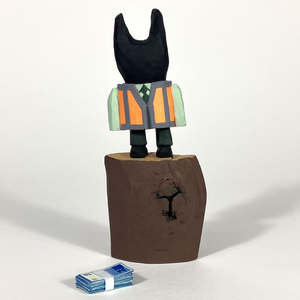 Cherry wood carved figure of a cat like humanoid form in a shirt and tie and a hi viz waistcoat. It’s stood on an uncarved section of the wood. A band of tiny paper printed banknotes is laying near the base of the figure.