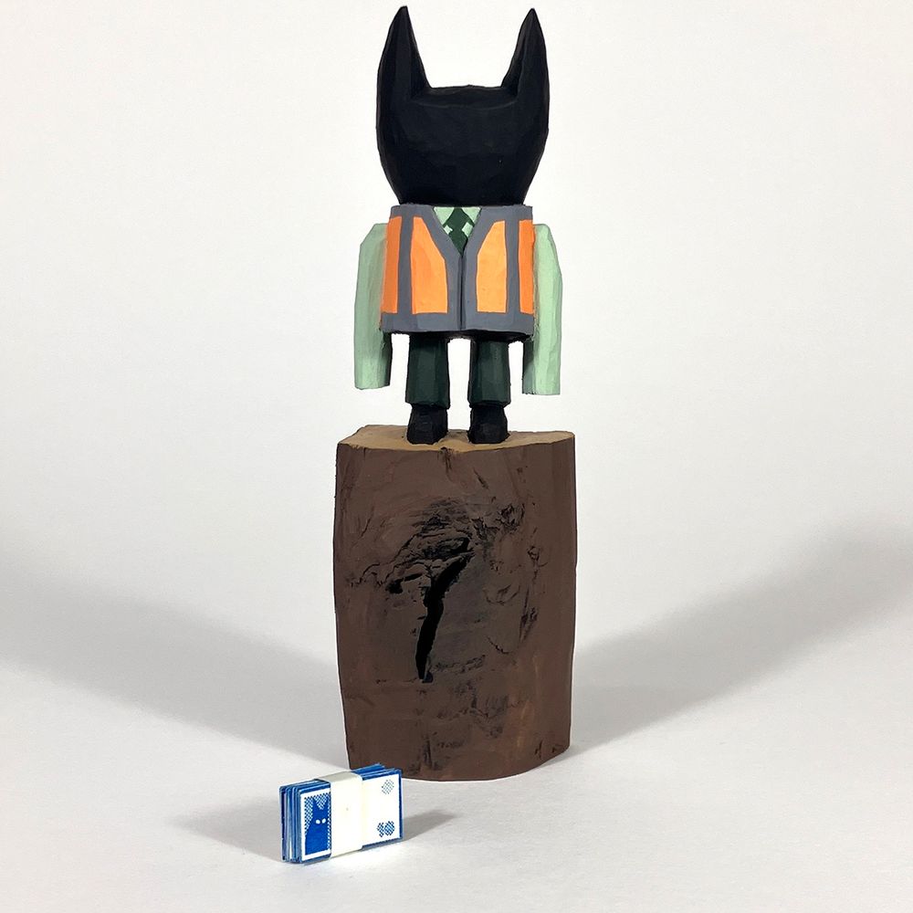 Cherry wood carved figure of a cat like humanoid form in a shirt and tie and a hi viz waistcoat. It’s stood on an uncarved section of the wood. A band of tiny paper printed banknotes is laying near the base of the figure