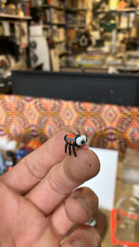 A life size house fly model kit by Modern Toss on the tip of a finger. The fly is cartoon like with big eyes and a hi viz vest painted on it. 