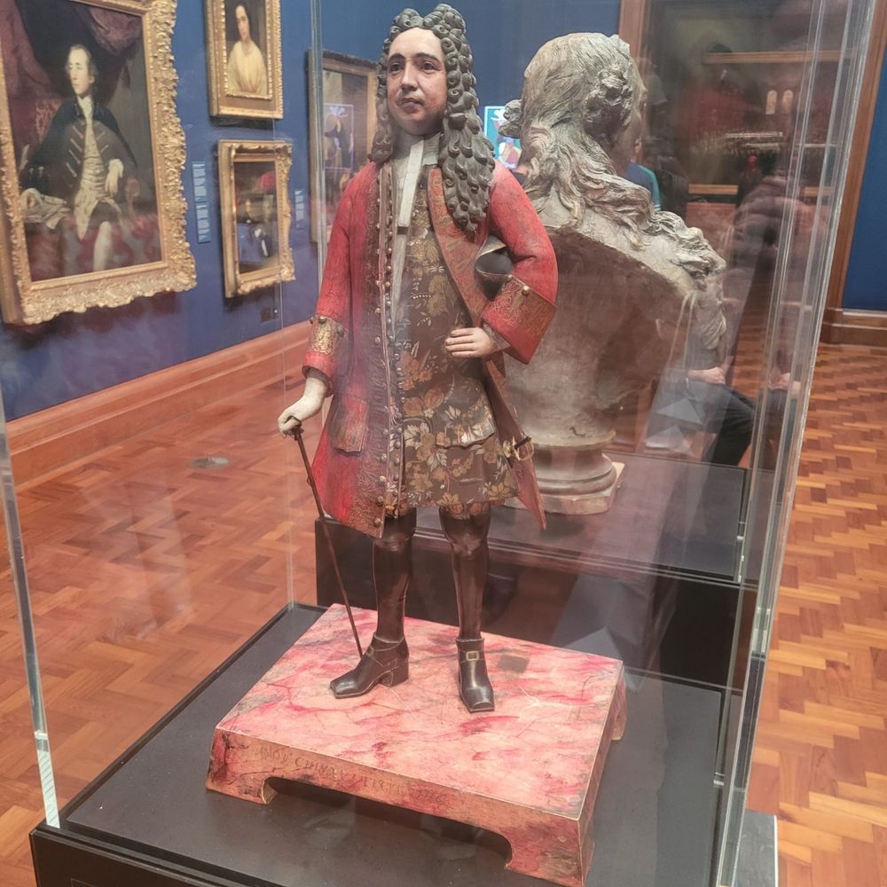 Photograph of a man in early-18th century clothing in a glass case at the National Portrait Gallery, London. 