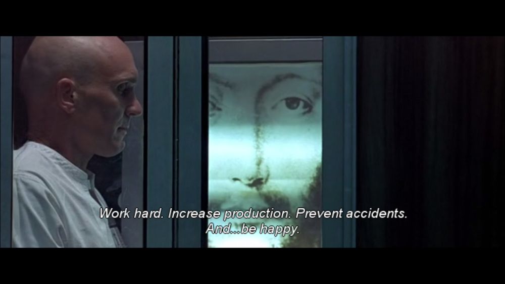 A still from a scene from the movie THX 1138 where the title character (Robert Duvall) visits a computerized confessional booth decorated with a portrait of Jesus (identified in the movie as “OMM 0000”); the captioning (from Jesus/OMM) reads: “Work hard. Increase production. Prevent accidents. And…be happy.”