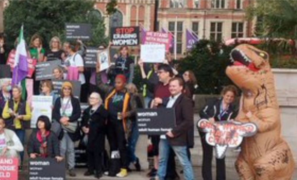 Close-up on the right half of the Bad Medical Taker's Xitter banner picture showing an anti-trans demonstration, people holding various signs including those of a purported dictionary definition of “woman” and also someone in a T. Rex costume