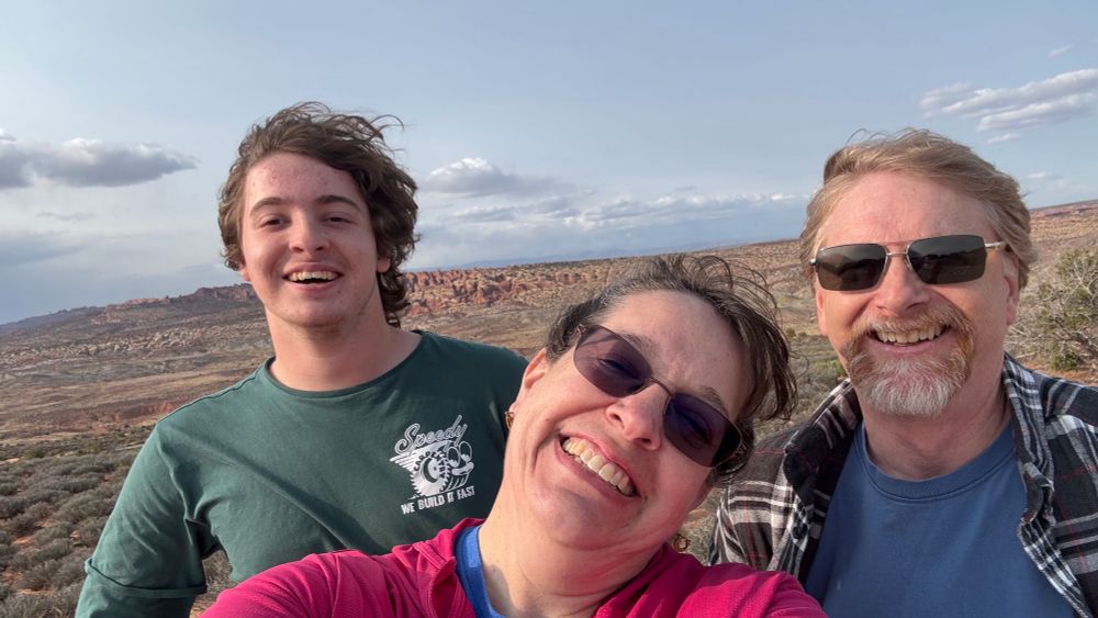 Three people in the vast open space of the desert southwest (Arches NP). All of us are wind blown and messy-haired but happy. 16 yo manchild, me, and husband 