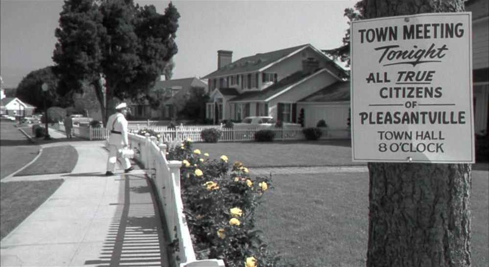 same image, zoomed out, showing a row of flowers that are color when everthing else is black and white. and a tree, with a sign on it that reads "town meeting. tonight. all true citizens of pleasentville, and here the word true is underlined, town hall. 8 o'clock. and now seeing its a scene from the film pleasentville. a film that seems like a cozy comedy, until the riots on the streets over, literal, color.
