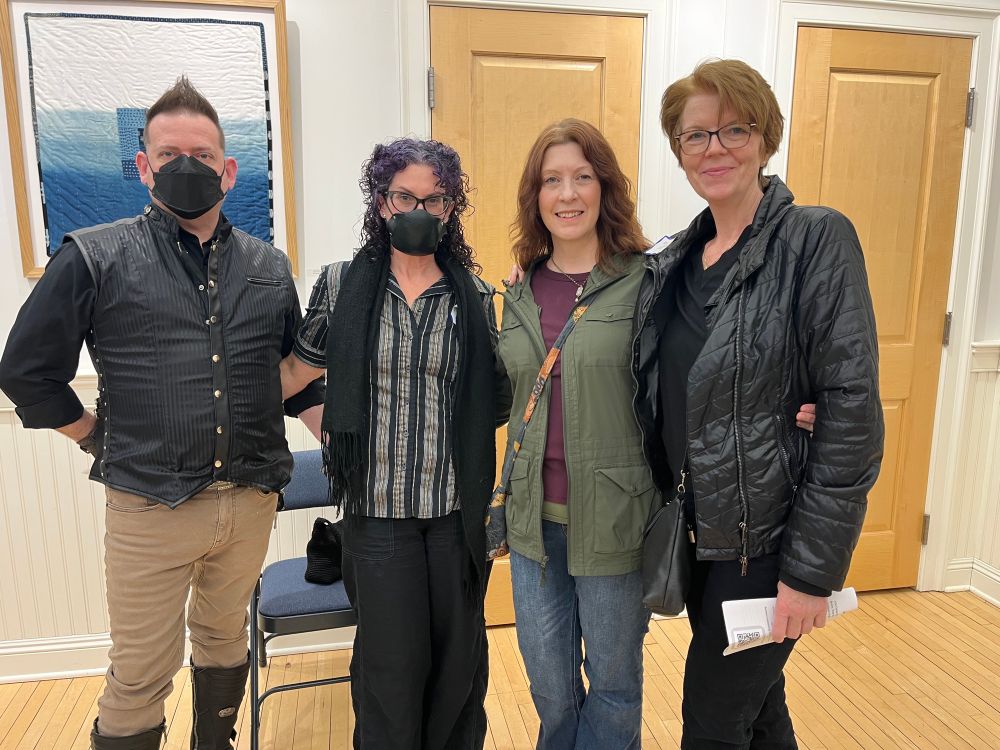 An image of artists friends standing in a line with arms around each other. 
Left to right: 
Joseph Marsocci with spiky dark hair, a black vest over black shirt, tan pants, tall black boots and black mask over his nose and mouth. 
Rachel Sclare with curly dark purple hair, black cats eye glasses, a black, silver, and gold striped button down shirt, black pants, and a black mask over nose and mouth. 
Gwyneth Amy with shoulder length auburn hair, an olive jacket over a raspberry shirt, and blue jeans.
Amy LaBossiere with short auburn hair, glasses, a black jacket over black shirt, and black pants- she holds the art show program in her left hand. 