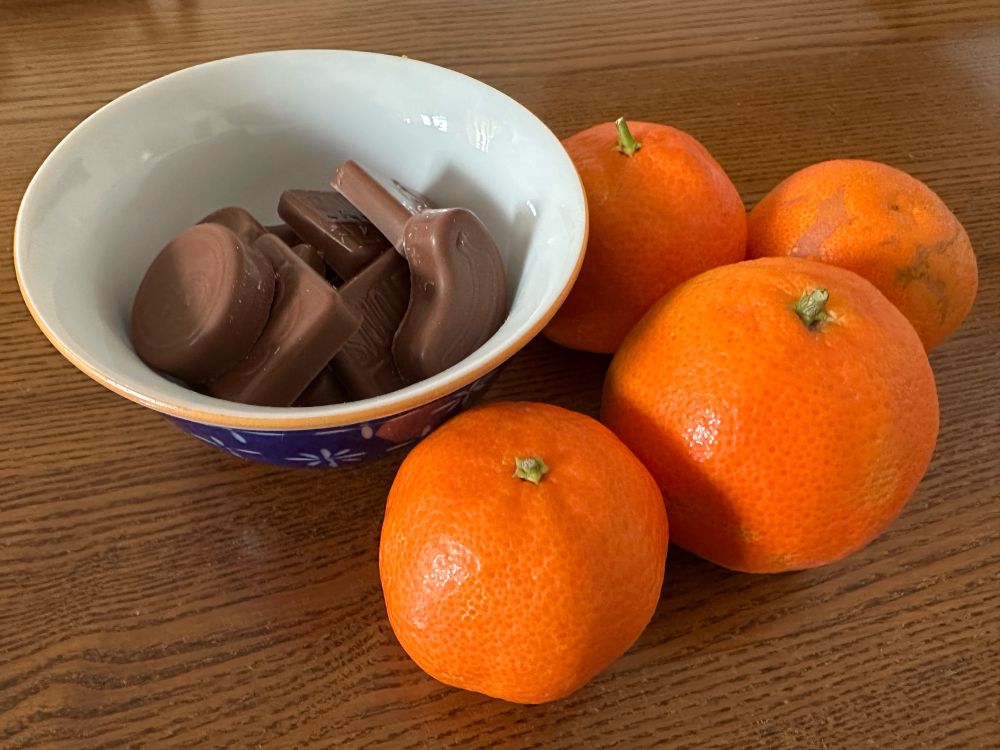 Una piccola ciotola di ceramica appoggiata su un tavolo di legno con alcuni cioccolatini all’interno, alla sua destra quattro clementine 