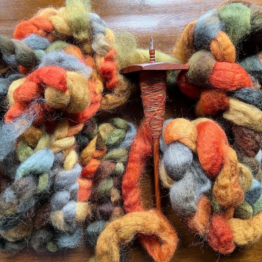 A pile of autumnal colored wool roving, with one big piece plus seven little pieces, with a wooden spindle holding the beginning of the handspun yarn. 