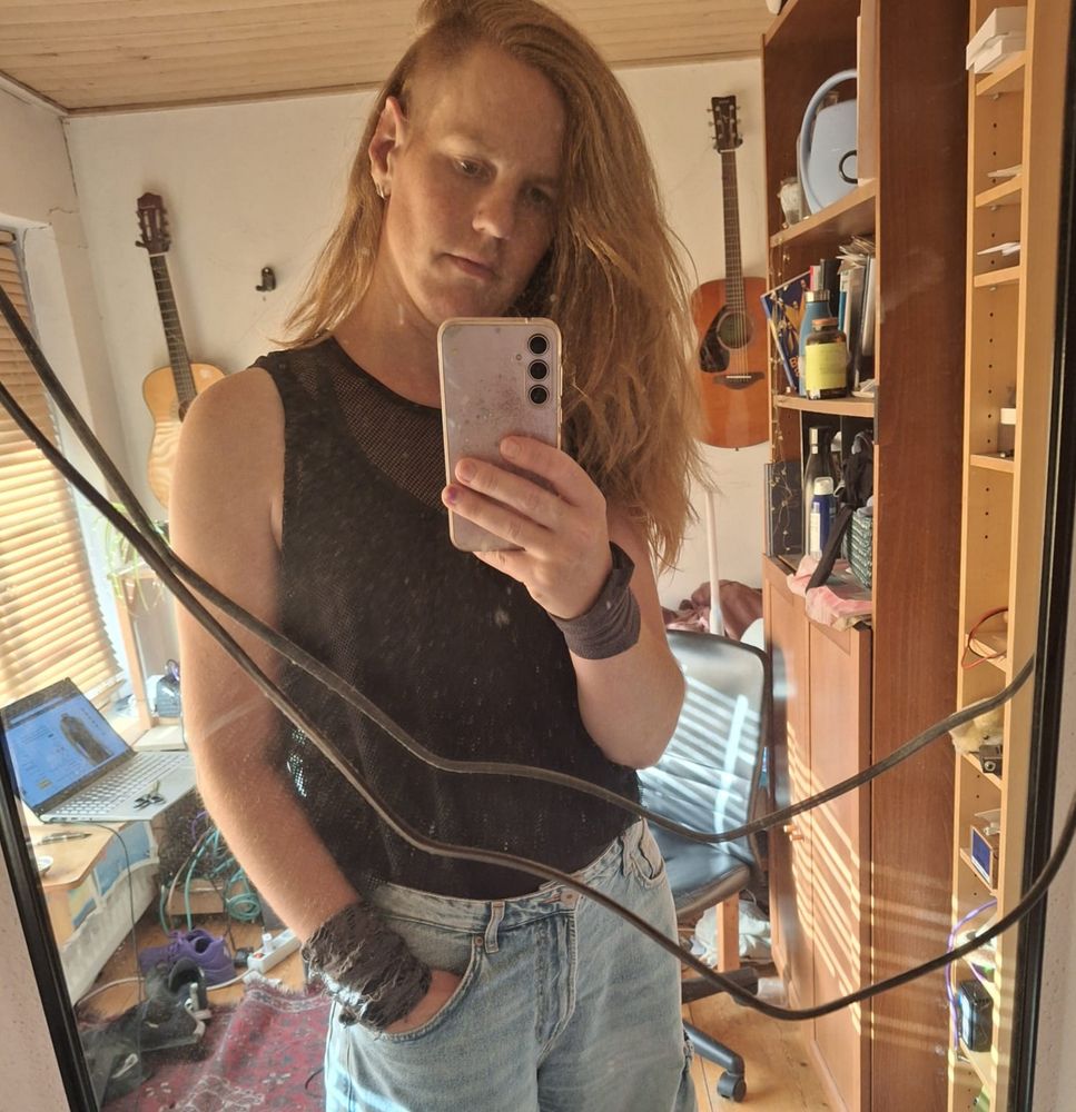 spiegelselfie van mij in een rommelige kamer. Ik heb lang roodbruin haar, met een opgeschoren zijkant. Ik draag een zwarte tanktop, een spijkerbroek, en armbanden