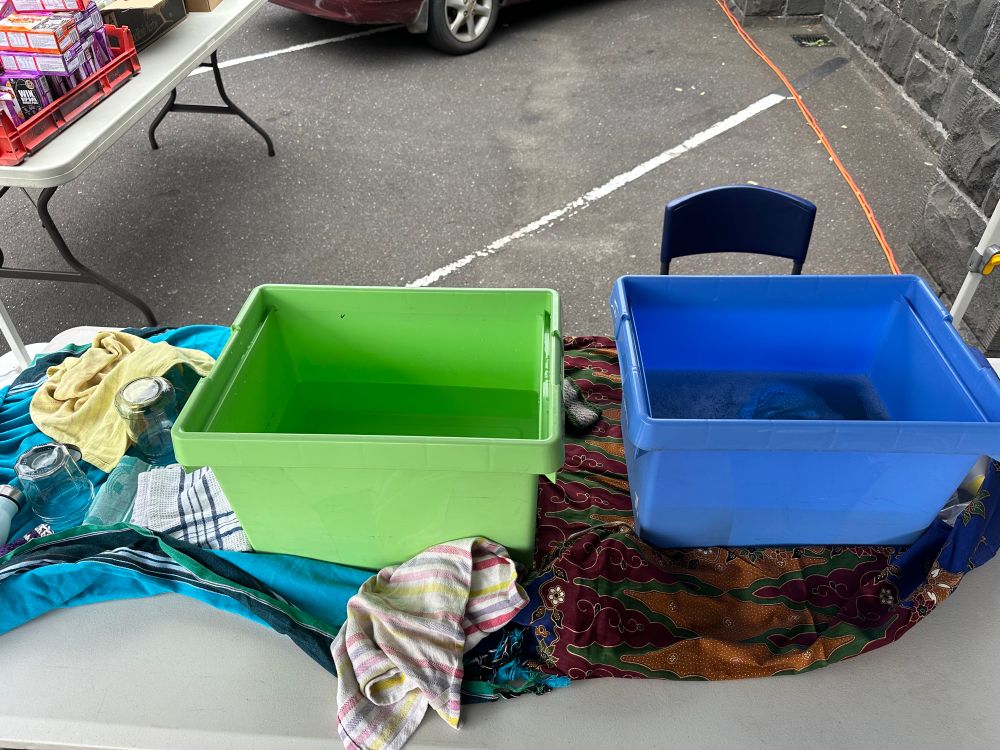Photo of a fold-out table with two buckets of water (one looks soapy) sitting on spme tea towels. with a chair behind one of the buckets.