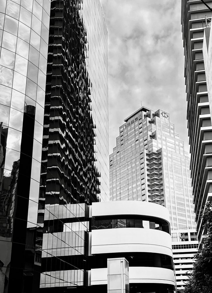 Black-and-white photo of tall modern buildings in Bangkok. A mirrored glass tower reflects wavy distortions, while a central skyscraper rises in the background.  