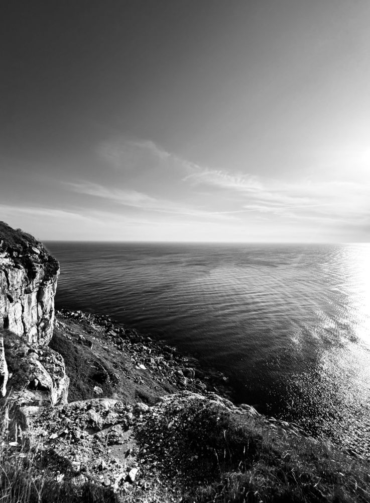 Standing at the edge of the cliffs at Portland, Dorset, looking out over a vast, silver-blue sea beneath a soft, clouded sky. The light is quiet and reflective, the horizon stretching endlessly.
