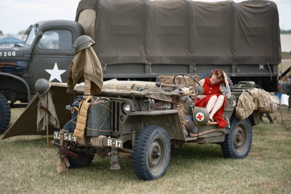 A woman in a red dress looks down to her lap. She is sitting on a Willis Jeep all in the style of the 1940’s