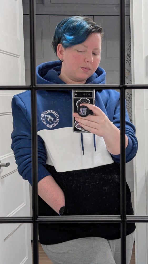 I'm wearing a new tricolor hoodie with horizontal color blocking of blue, white, and black. My right hand holds my phone with a pop socket and my left hand is in the hoodie pocket. I am wearing light grey sweatpants. The photo is taken standing in a mirror with decorative black window muntins. An open doorway is behind me.