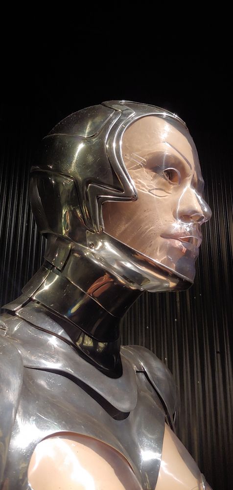 Detail view of the face of the chrome armor by Mugler worn by a mannequin. Plexiglass covers the face with small holes at the eyes, nose, and mouth. The chest is seen through plexiglass as well. Chrome armor covers the side and back of the head up to the cheek bone. The armor is layered to articulate on the neck.
