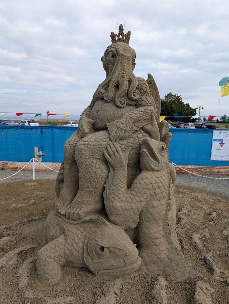 The elephantine figure of Cthulhu and his octopoid head, sculpted in sand. He is attended by a couple of fish-headed mer people that rise out of the sand to form his throne. 