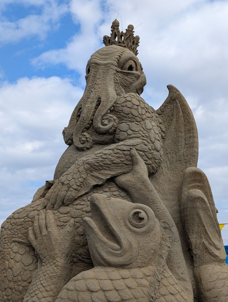 A close up of Cthulhu, sculpted in sand. He bears a crown and one of his fish-person attendants supports his finned arm with their hand. Their wide fish mouth hangs open as their round eyes gaze skyward. 