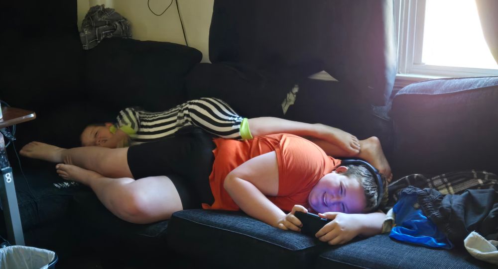 Two children stacked on top of each other laterally head to feet. The top one is in Beetlejuice body suit pajamas. The lower one is in an orange t and black shorts. The sun glares down on them through parted curtains. 