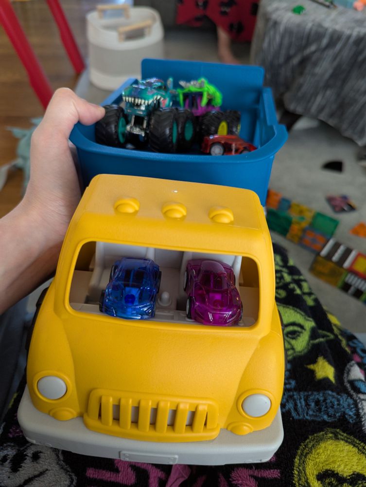 A yellow dump truck with a blue truck part. In the truck part are 2 toy monster trucks and a little red car. In the seats of the dump truck there is a small purple and blue car. 