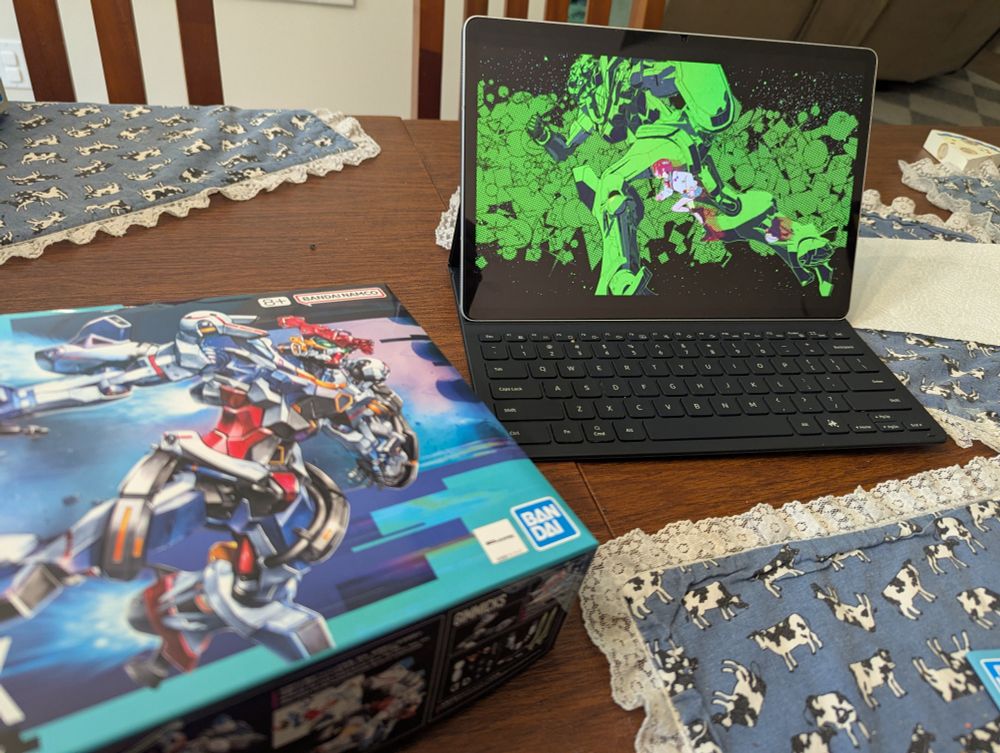 A tablet playing an episode of Gundam GQuuuuuux along with the lid of a GQuuuuuux model kit. 