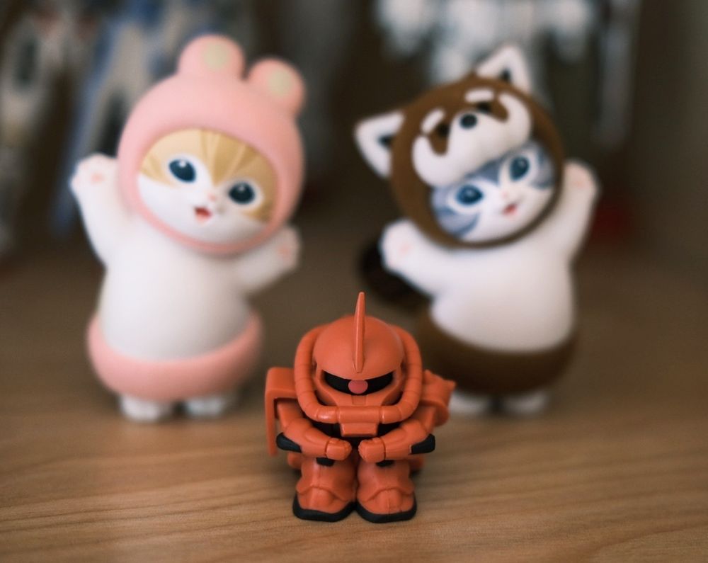 A small red zaku sitting down with it's arms around its legs looking sad with a dancing car wearing animal costumes on either side. 