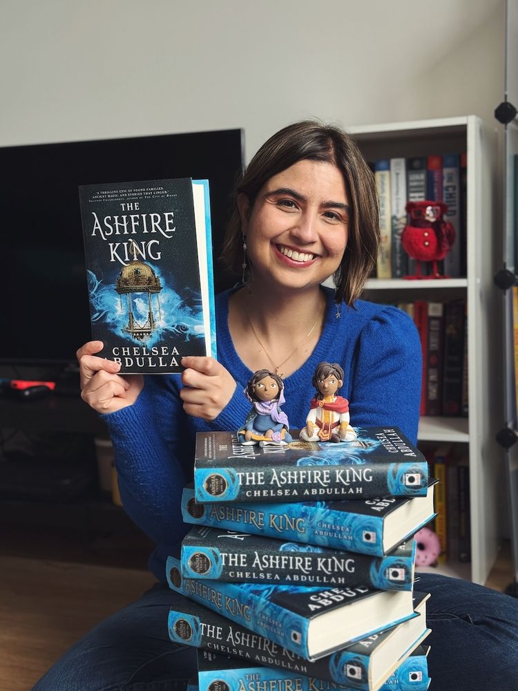 A photo of me, Chelsea, holding up a copy of my newly released book, THE ASHFIRE KING, behind a stack of ASHFIRE KING copies, all spine out. Two clay figurines of Loulie and Mazen sit atop the stack.