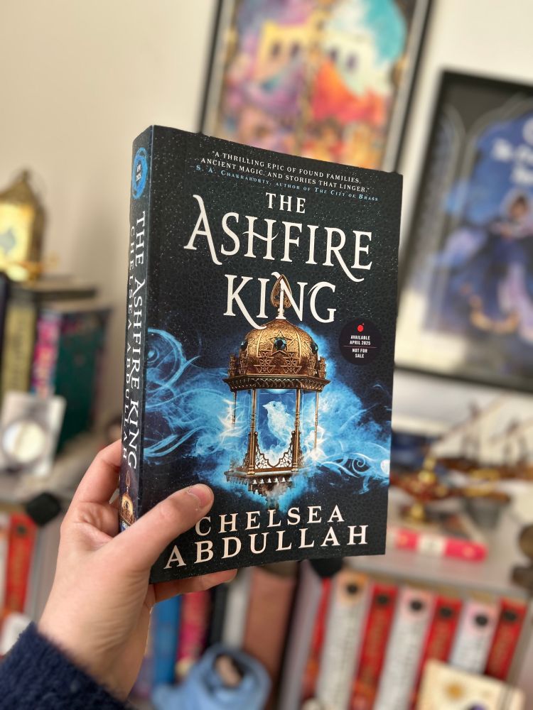 A photo of an ARC of THE ASHFIRE KING against the backdrop of my bookshelves, which are filled with copies of THE STARDUST THIEF 