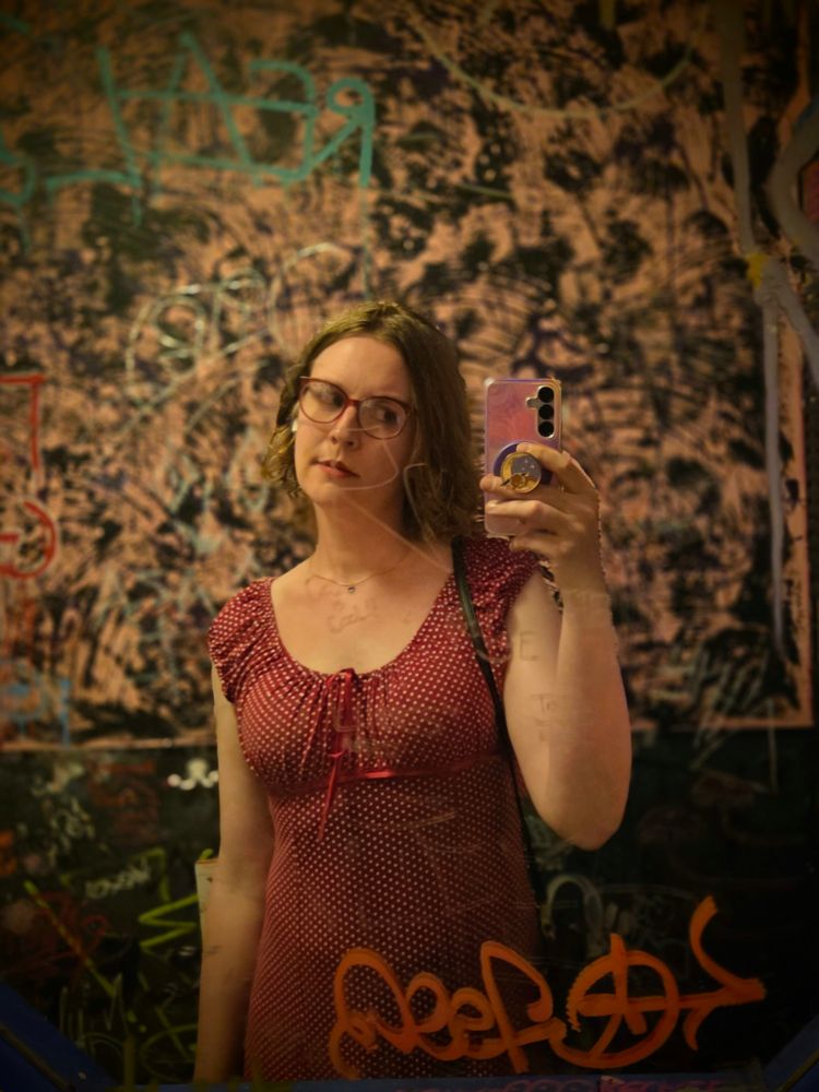 a trans woman talking a selfie in a women's bathroom covered in graffiti, she's wearing a red polka dot dress and red glasses