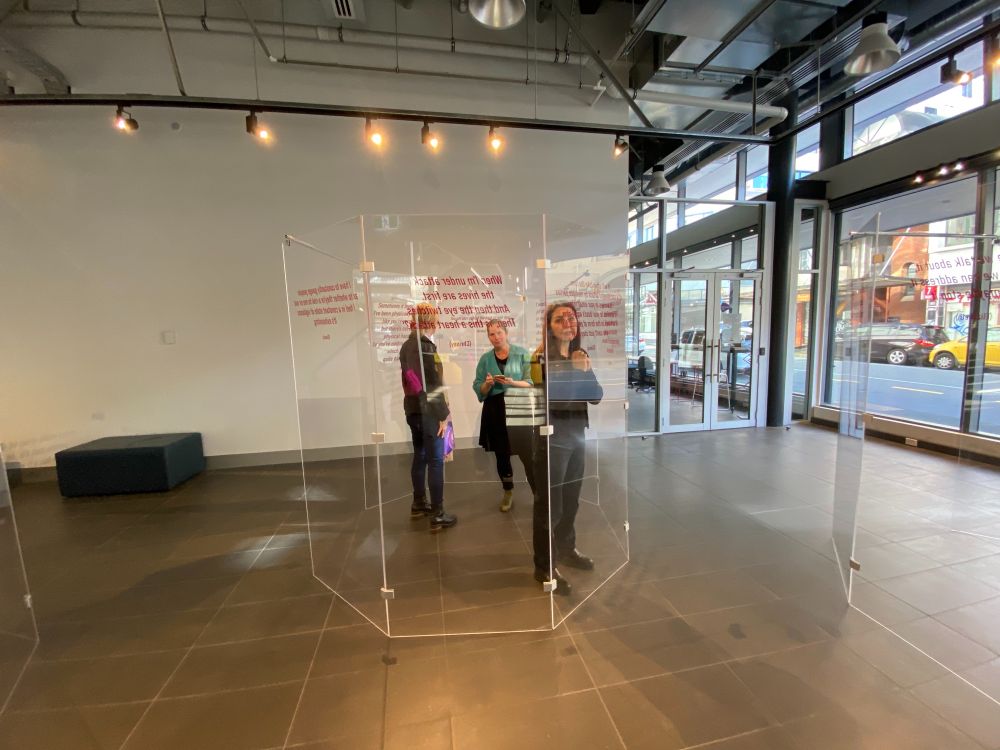 3 women inside a glass cage Reading words printed on the glass of women's experiences of online misogyny