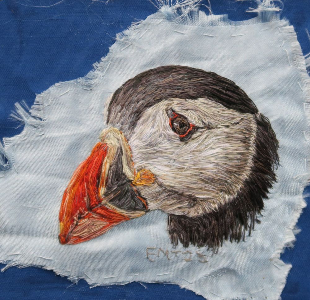 A royal blue fabric background with a sandpapered edge light blue sample of fabric in the middle which has a hand embroidered Puffin head in the middle - the head is turned to the left.