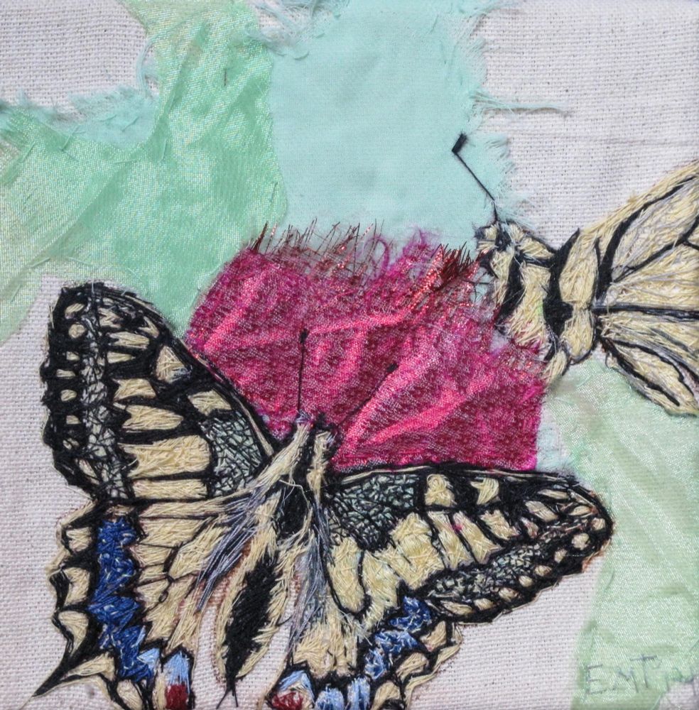 A swallowtail butterfly with its wings open which is hand embroidered, next to another hand embroidered body of  a swallowtail. Both are on a pink fabric thistle on a cream and green fabric background 