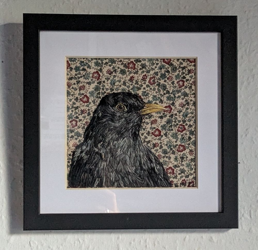 A hand embroidered male blackbird head to mid chest on an autumnal floral and vine patterned fabric background. Set in a white mount and black frame, seen on a light grey wall 