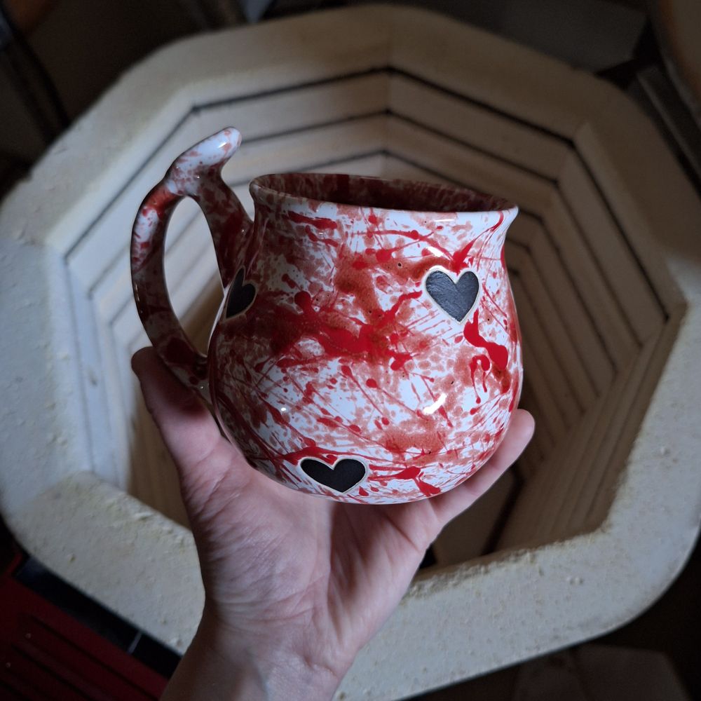 My left hand holding the mug I just pulled from the kiln in the background. The curvy mug feature a glossy white background with lots of red splatters and matte black hearts. ❤️🖤