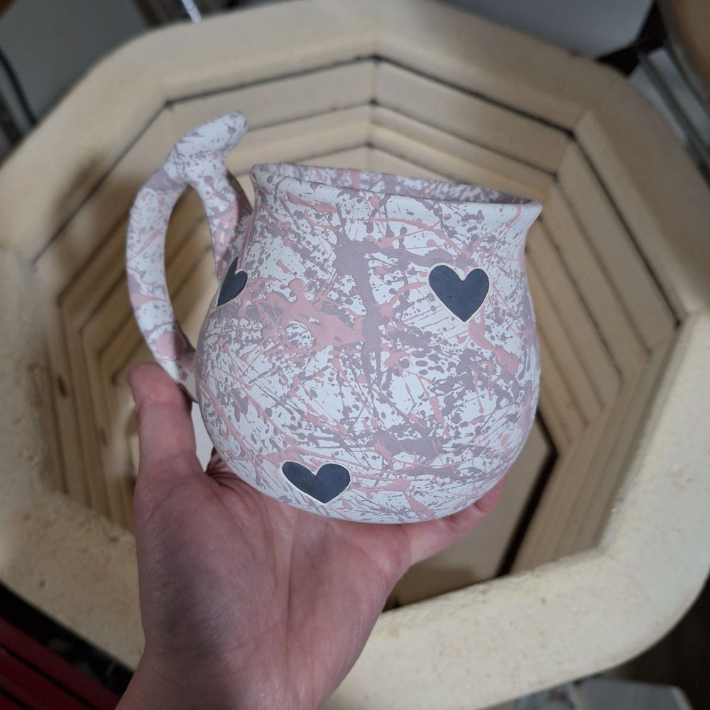 My left hand holding a glazed mug before placing it into the kiln (featured behind the mug) to be fired. The mug is curvy with a handle that has a thumb rest, and is decorated with black hearts surrounded by a field of pale red splatters on a white background.