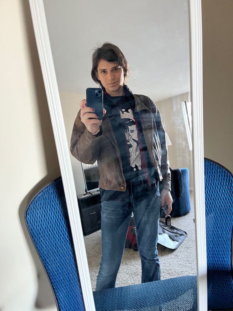 me in a leather jacket and jeans. my hair is pulled back. i look severe; but one corner of my mouth is turned up. my right hand is taking a selfie and the left is holding my car keys. in the background is a tv and shaded window.