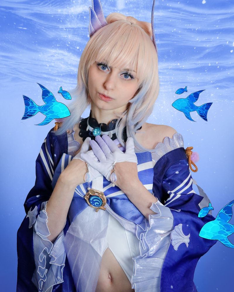 Cosplay of Kokomi from Genshin Impact, featuring a blue and white outfit with flowing sleeves, a necklace with a blue pendant, and edited decorative fish and water elements in the background. The cosplayer is putting both hands on the chest, which could express varied emotions including gratitude and relief.