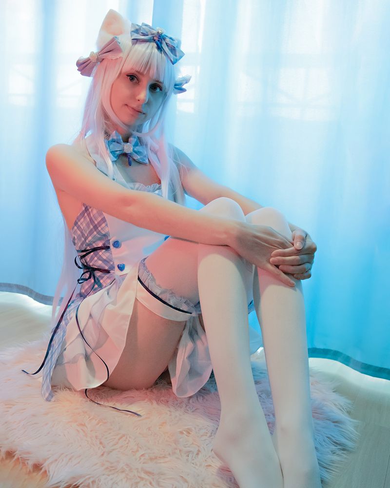 Cosplay of Vanilla from Nekopara with long white hair wearing a blue and white checkered dress with bows, sitting on a fluffy rug in front of a blue curtain.