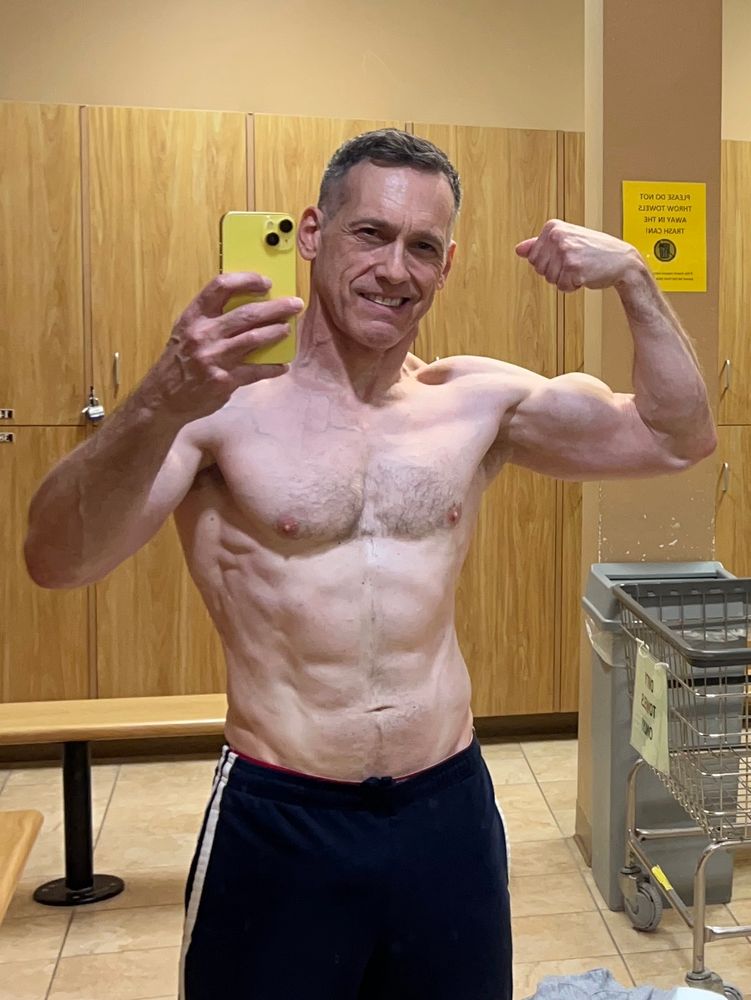 A mirror selfie of myself shirtless in the gym locker room. I’m flexing my left arm in a bicep curl pose.