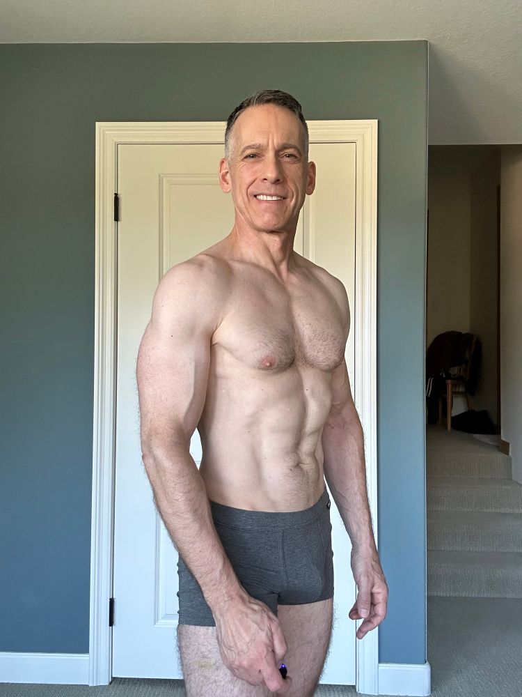 A selfie I took (in undershorts) documenting my bodybuilding/cutting progress. Similar to a previous image where I’m in a three-quarter-ish turn but showing more definition in my torso. I’m happy and smiling.