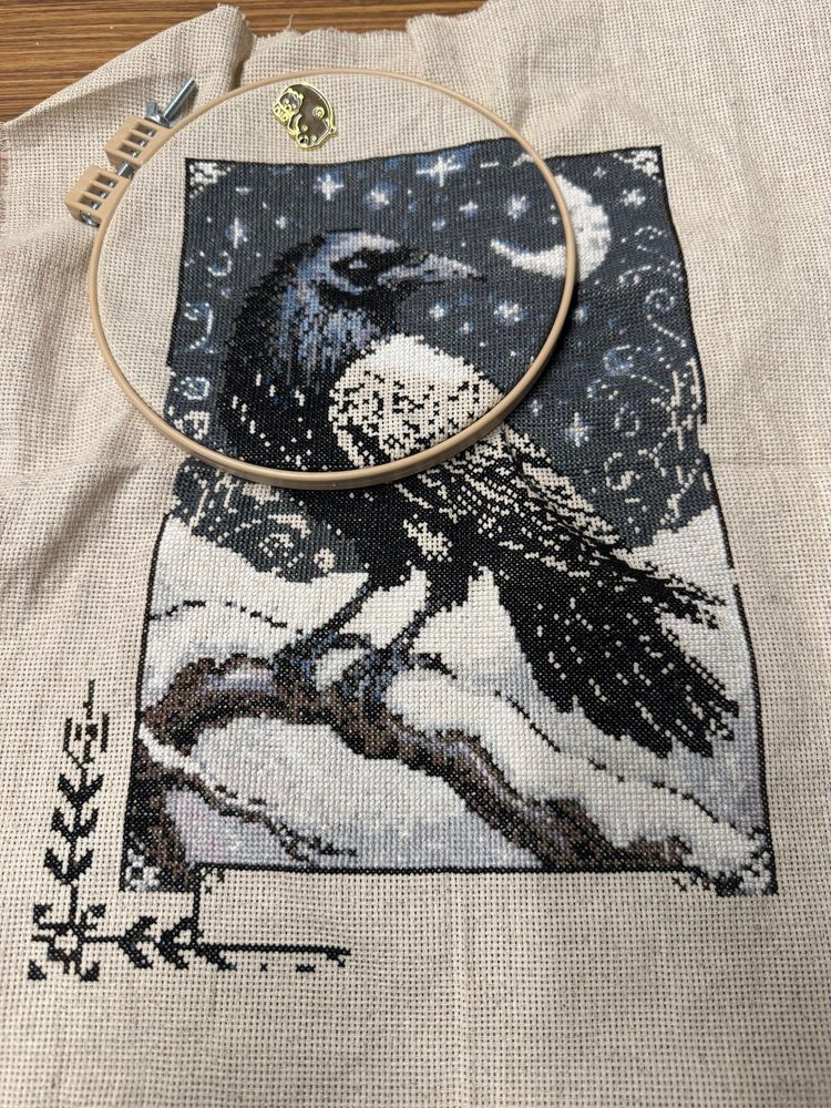 In progress cross stitch of a raven standing on a snow-covered branch. Its wing, and part of the background, still need to be filled in.