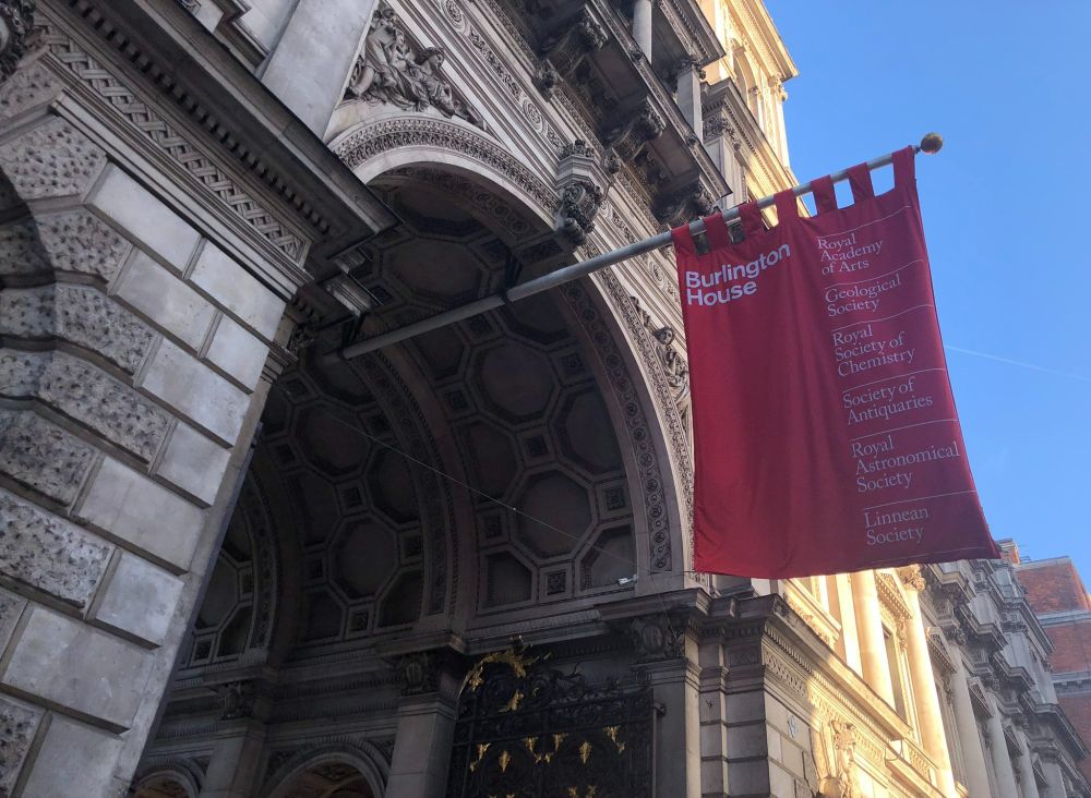 An image of the entrance to the courtyard at Burlington House; a large, ornate stone arch with black iron gates below adorned with gold details. A red banner hangs overhead, reading "Burlington House, Royal Academy of Arts, Geological Society, Royal Society of Chemistry, Royal Astronomical Society, Linnean Society". It's a sunny day with clear blue skies behind, and sunlight illuminating the walls behind the banner.